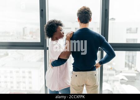 Une femme africaine positive embrasse avec son beau ami brunette européen, se détendre à la maison, se donner chaud câlin les uns aux autres, se tenir près de la fenêtre arrière-groupe Banque D'Images