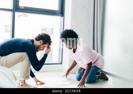 Des partenaires multiculturels spécialisés dans les petites entreprises développent un projet futur, s'agenouillant sur le sol dans la salle de séjour, regardant les papiers, pensant à la façon de me faire Banque D'Images
