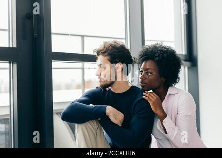 Heureux couple interracial à à la fenêtre de leur nouvelle maison louée, assis dans la salle vide. Déménagement en nouvelle house concept Banque D'Images