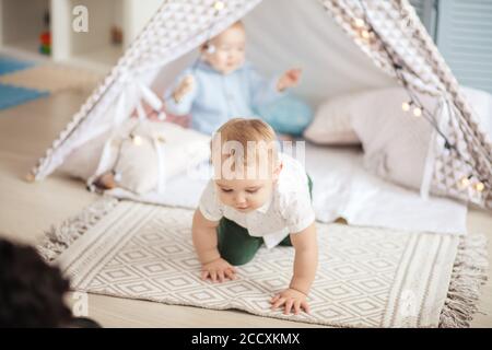 Deux jeunes enfants du même âge s'assoient sur un tapis dans une tente à tipi, s'amusent et jouent avec des guirlandes de Noël dans une chambre confortable. Banque D'Images