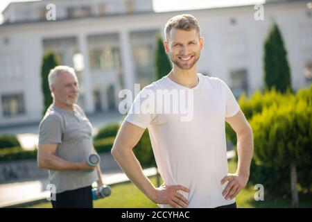 Homme barbu souriant, mains sur les hanches, homme aux cheveux gris tenant des haltères derrière Banque D'Images