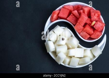 La pastèque et le melon mûrs, coupés en cubes, sont disposés sur un plat de yin-yang sur fond noir. Fruits d'été juteux et mûrs. Lumière vive. Copier l'espace. Banque D'Images