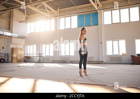 Gracieuse grande fille de gymnaste se tient avec dos à l'appareil photo, au milieu de la salle de sport, porte de belles leggins noirs et soutien-gorge, prêt à commencer l'entraînement Banque D'Images