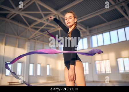 Charmante belle gymnaste d'art lève la main, agitant avec long ruban violet, regardant directement l'appareil photo, tourné de dessous, concept de sport professionnel Banque D'Images