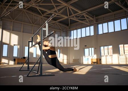 Une jeune athlète féminine attirante est accroché à la barre de ballet, met la jambe en place, tient la barre métallique, ferme les yeux avec une expression agréable, appréciant le temps de l'entraînement roo Banque D'Images