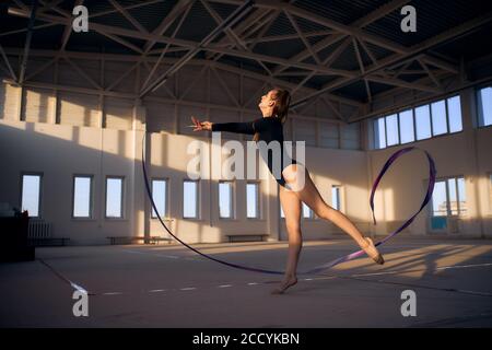 Belle fille gracieuse vêtue de vêtements de sport noirs, élément de gymnastique artistique, tenant le ruban violet, s'agiter, regardant loin, coup de pied, p Banque D'Images