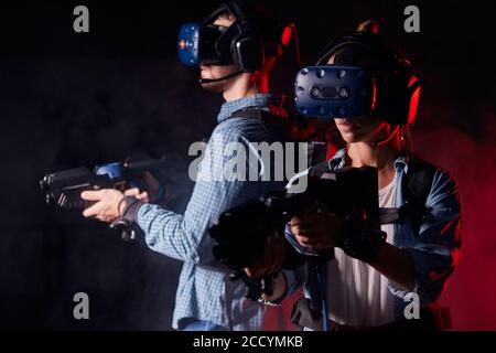 Homme et femme avec des pistolets vr et des micro-casques sur fond noir isolé avec rétroéclairage au néon rouge. Divertissement, espace de copie Banque D'Images