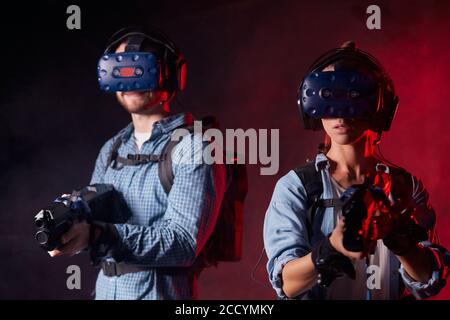 Homme et femme avec des pistolets vr et des micro-casques sur fond noir isolé avec rétroéclairage au néon rouge. Divertissement, espace de copie Banque D'Images