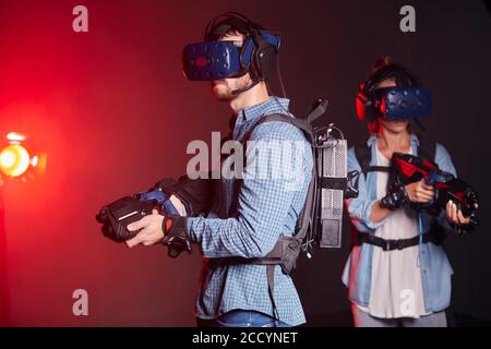 Homme et femme avec des pistolets vr et des micro-casques sur fond noir isolé avec rétroéclairage au néon rouge. Divertissement, espace de copie Banque D'Images