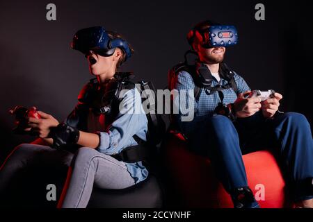 Un jeune homme et une jeune femme jouent au jeu vidéo assis sur un canapé isolé sur fond noir, rétro-éclairé au néon. À l'aide de manettes, porter des casques, des lunettes de protection Banque D'Images