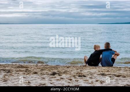 Deux frères se lient sur la beauté de l'océan comme ils s'assoient sur la plage, avec le frère cadet réconfortant l'ancien avec un bras autour de lui. Banque D'Images