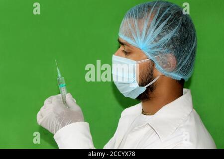 Jeune médecin avec seringue, homme dans le masque médical se préparant à l'injection sur fond vert, concept de vaccination pendant la pandémie de Covid-19. Banque D'Images