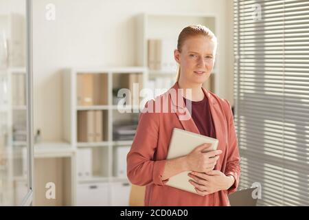Portrait à la taille haute d'une jeune femme d'affaires aux cheveux rouges regardant l'appareil photo tout en se tenant sur le lieu de travail dans un bureau éclairé par le soleil, espace de copie Banque D'Images