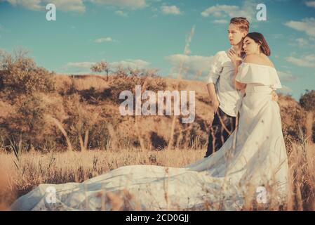 jeune mariage couple d'un gars et d'une fille, dans une robe blanche, debout dans un champ, sur la toile de fond d'un ciel bleu nuageux, coucher de soleil. Amour du jeune peop Banque D'Images