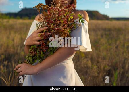 portrait d'une belle fille en gros plan. Il tient un bouquet de fleurs sauvages dans ses mains. Dans une robe blanche. Sur fond de ciel bleu nuageux. Ha Banque D'Images