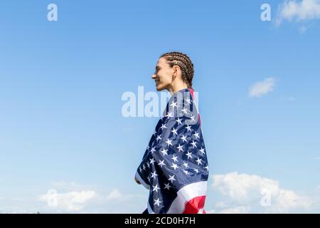 Bonne jeune femme américaine avec des afro-braides enveloppés dans des rayures et étoiles drapeau USA contre le ciel bleu. Banque D'Images