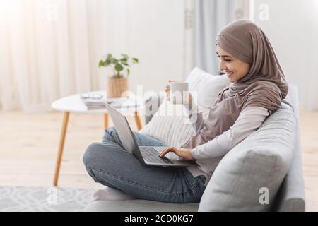 Détente à l'intérieur. Femme musulmane souriante utilisant un ordinateur portable et prenant du thé à la maison Banque D'Images