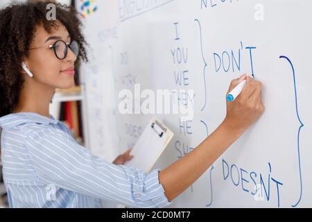 Professeur en ligne. Fille afro-américaine en lunettes avec casque sans fil écrit avec marqueur sur tableau noir Banque D'Images