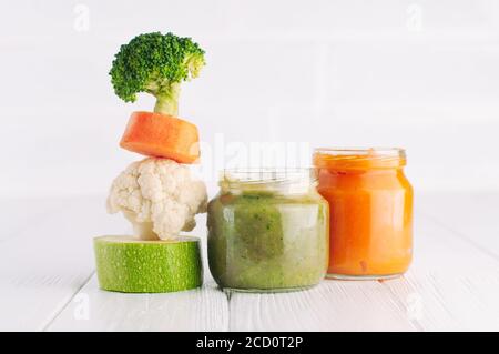 Concept de nourriture naturelle pour bébé. Purée de légumes dans deux bocaux près de la pyramide de brocolli, carottes, chou-fleur et courgettes sur fond blanc avec du blan Banque D'Images