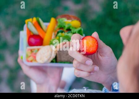 Concept d'alimentation saine : jeune femme mangeant à partir d'une boîte à lunch remplie de sandwich, de pain croustillant, de fruits et de légumes à l'extérieur; concentration sélective sur la prune Banque D'Images