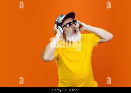 Danse folle. Portrait de l'homme hipster senior utilisant des appareils, gadgets isolés sur fond orange studio. Concept de mode de vie technologique et joyeux pour les personnes âgées. Couleurs tendance, toujours jeune. CopySpace pour votre annonce. Banque D'Images