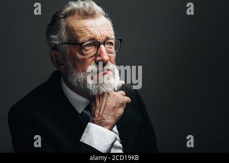 Écrivain barbu mature en lunettes et yeux expérimentés confiants, vêtu d'un costume habillé noir pensant à la main sous le menton posant isolé sur le noir Banque D'Images