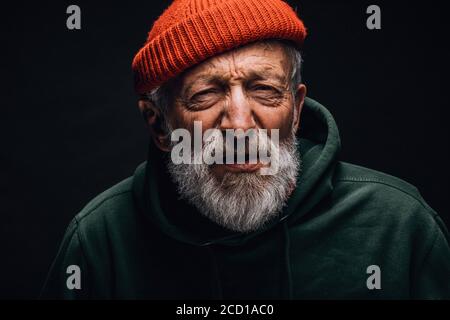 Vieux sans-abri vêtu de vêtements d'extérieur usés mais propres et chapeau regardant la caméra avec les yeux vissés et expression interrogative ayant vue ou Banque D'Images