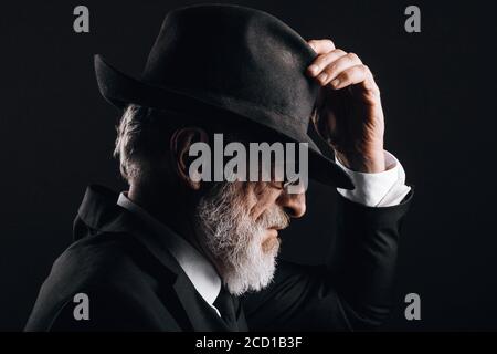 Portrait de gentleman âgé élégamment habillé de style classique, chapeau trilby et costume noir. Mode masculin, look classique. Studio tourné dans l'obscurité Banque D'Images