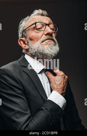 Homme d'affaires à barbe mature portant des lunettes en costume habillé noir réglable son cravate sur fond noir Banque D'Images