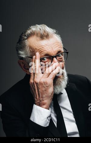 Homme politique à barbe mature en lunettes et yeux expérimentés confiants, vêtu d'un costume habillé noir pensant avec la main sous le menton posant isolé sur b Banque D'Images