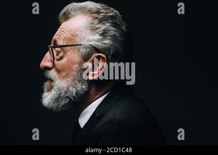 Profil de la tête de l'intelligent barbu gris cheveux scientifique écoute rapport, vêtu d'un costume noir habillé, assis isolé au-dessus de l'obscurité Banque D'Images