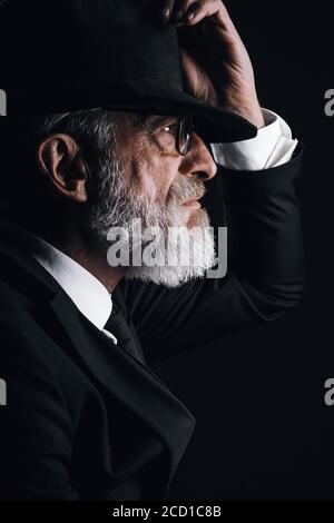 Homme barbu d'âge ancien, à l'image d'un agent secret anglais portant un costume noir avec chapeau sur les yeux posé sur un fond sombre. Banque D'Images