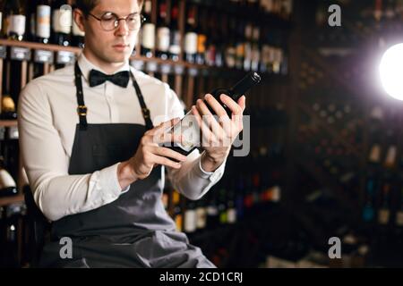 Jeune caviste caucasien vêtu d'une chemise blanche et d'un noeud papillon dans une grande boutique de vins présentant une bouteille de vin rouge au client Banque D'Images