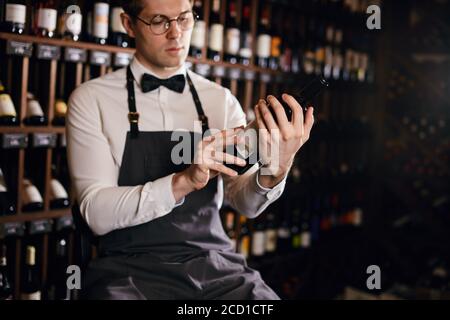 Beau et jeune sommelier brun souriant avec une bouteille de vin sur le fond de la maison de vin sombre avec étagères de bouteilles avec alcool Banque D'Images