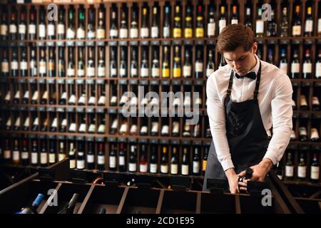 Beau et jeune sommelier brun souriant avec une bouteille de vin sur le fond de la maison de vin sombre avec étagères de bouteilles avec alcool Banque D'Images