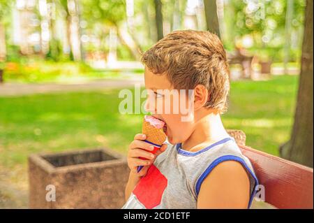 Joli jeune garçon dans un t-shirt gris est assis sur un banc dans le parc lors d'une journée ensoleillée et manger un cône de crème glacée Banque D'Images