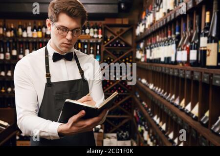 Sommelier professionnel de sexe masculin habillé de noeud papillon et de tablier écrit au bloc-notes, mettant en place des informations sur les bouteilles de vin dans la maison de vin Banque D'Images