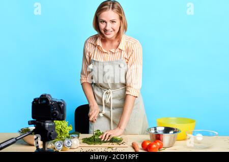 Belle femme blonde souriante se tient devant une table en bois pleine de légumes frais et ingrédients biologiques pour cuisiner des repas sains, couper Banque D'Images