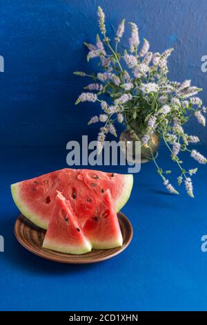 Trois tranches de pastèque fraîche mûre sont dans une assiette en argile bleu foncé. Dans un vase, un bouquet d'origan. Verticale Banque D'Images