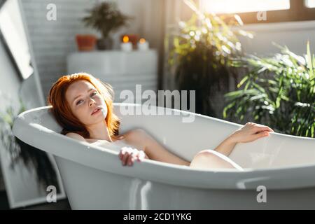 Jolie jeune femme aux cheveux rouges se détendant dans la baignoire de l'hôtel Spa, profitant de la tranquillité qui combine des éléments de design harmonieux avec premi Banque D'Images