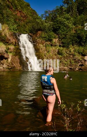 Femme en maillot de bain, portant un gilet de sauvetage, barboter dans l'eau pour nager sous une chute d'eau de 45 mètres, Waimea Valley, Oahu Island, Haleiwa, Hawaii, États-Unis Banque D'Images