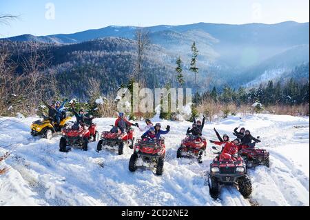 Yaremche, Ukraine - 02 février 2020 : des voyageurs heureux se levant les mains et souriant en conduisant des quads sur une colline enneigée avec de belles montagnes en arrière-plan. Concept de voyage et de quad. Banque D'Images