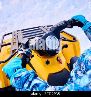 Gros plan des mains des femmes dans des moufles sport imperméables bleues conduisant un véhicule tout-terrain. Femme pilote tenant le guidon du quad jaune avec de la neige blanche sur fond. Concept de quad. Banque D'Images