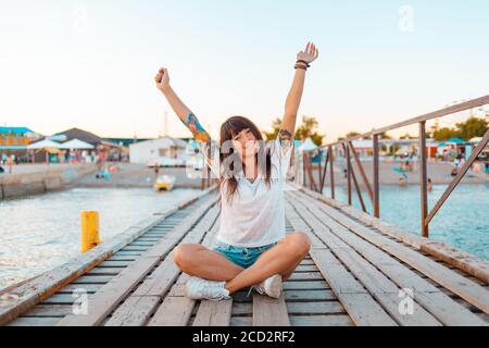 Une jolie femme avec des tatouages est assise à pieds croisés sur une jetée en bois au bord de la mer, ses mains soulevées dans la joie. Le concept des vacances d'été. Banque D'Images