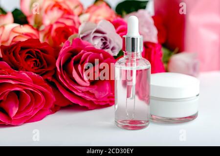 Bouteille cosmétique en verre, crème, sérum, huile sur fond floral de table blanc. Fleur rouge roses rose naturel produit de beauté biologique. Spa, soins de la peau, bain Banque D'Images