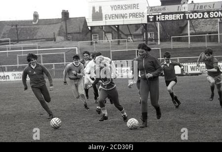 Années 1980, histoire, deux filles en costume de sport qui ont un coup d'envoi sur le terrain de football du York City FC sont pourchassés par des garçons, tous les jeunes qui travaillent sur le terrain ayant un peu de plaisir avant le match. Les terrasses de football de salon étaient les spectateurs debout et regarder un match peut être vu. Banque D'Images