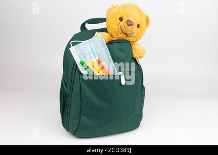 Sac à dos vert avec un ours en peluche et des marqueurs et masque de protection dans la poche avant Banque D'Images