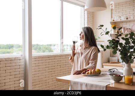 Image d'une jolie femme mûre debout à l'intérieur de la cuisine à la maison, boire du vin Banque D'Images