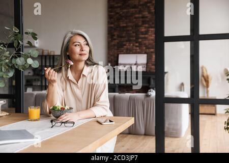 Photo d'une femme adulte étonnante assise dans la cuisine à l'intérieur à la maison tout en mangeant de la salade. Banque D'Images