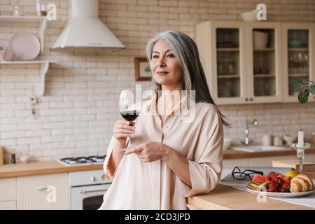 Photo d'une jolie femme mûre à l'intérieur de la cuisine à la maison, boire du vin Banque D'Images